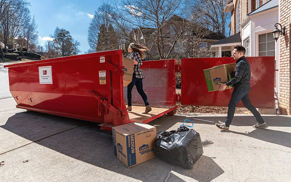 Two people carrying boxes into a roll-off dumpster in the driveway.